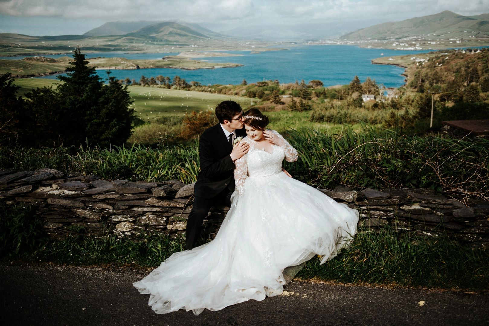 Groom kisses his bride, both of them sitting on a retaining wall with green Irish landscapes in the background