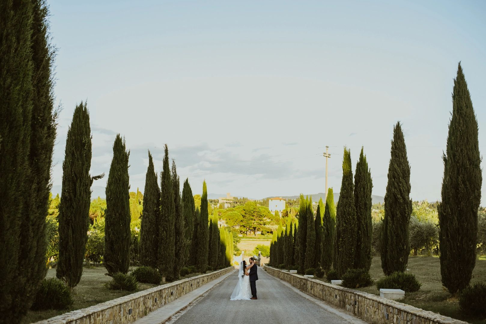Bride and groom walking among evergreen trees lining a driveway of a wedding venue in Italy