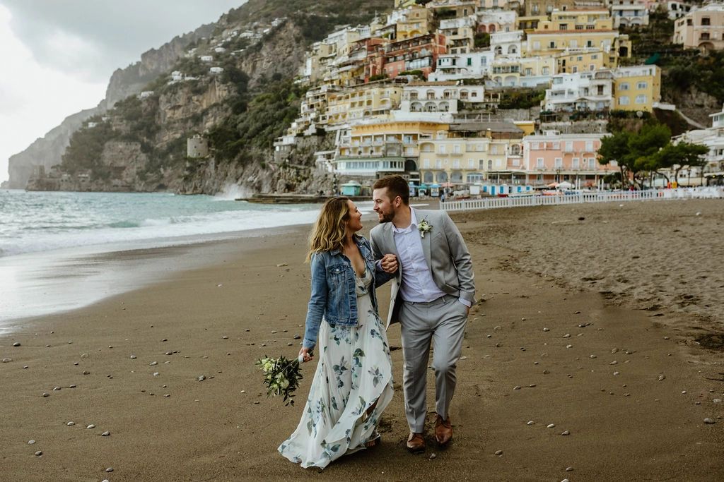 A couple walking along the beach with a few Amalfi buildings in the background during their elopement in Italy