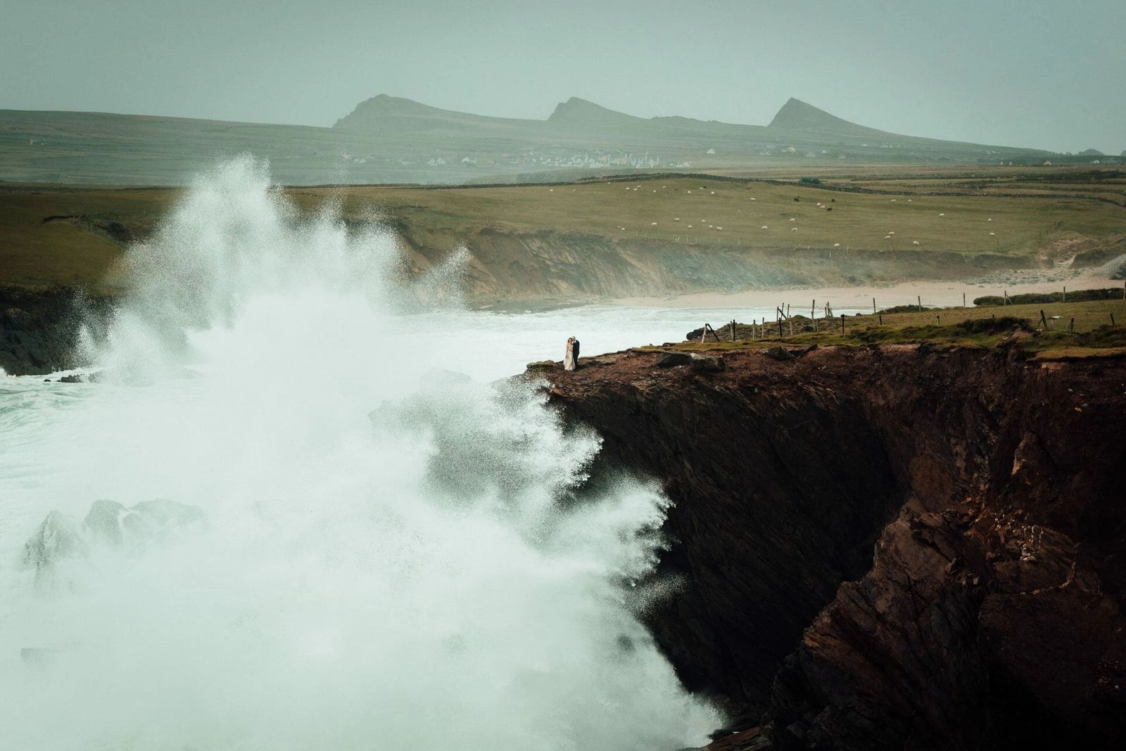 A couple stand at the cliff edge as a huge wave bursts against the rocks, Clogher Strand and the Three Sisters peaks behind them.