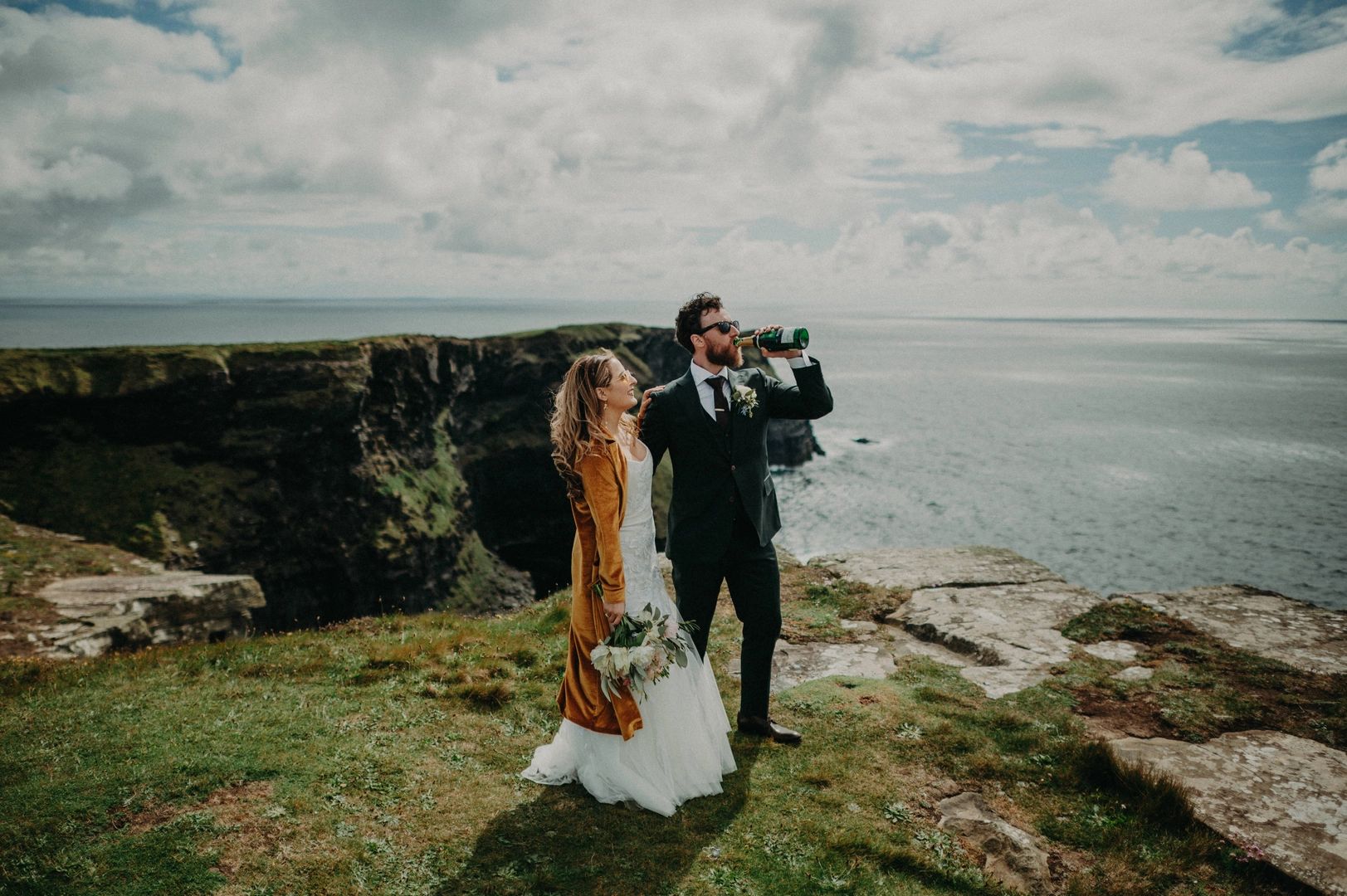 Bride and groom atop Hag’s Head during their elopement in Ireland, with the groom drinking a bottle of wine