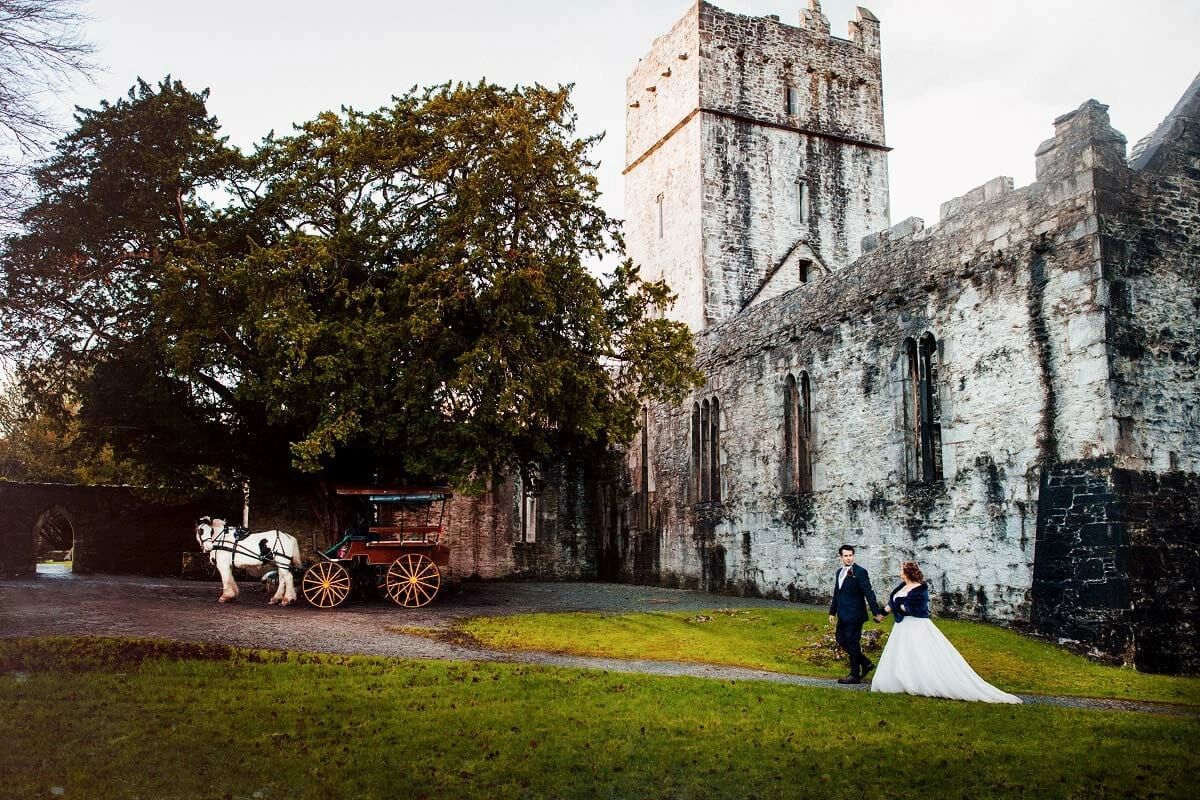 Monastery ruins in the background, a large tree in front, a horse carraige, and the newlyweds walking during their destination wedding in Ireland
