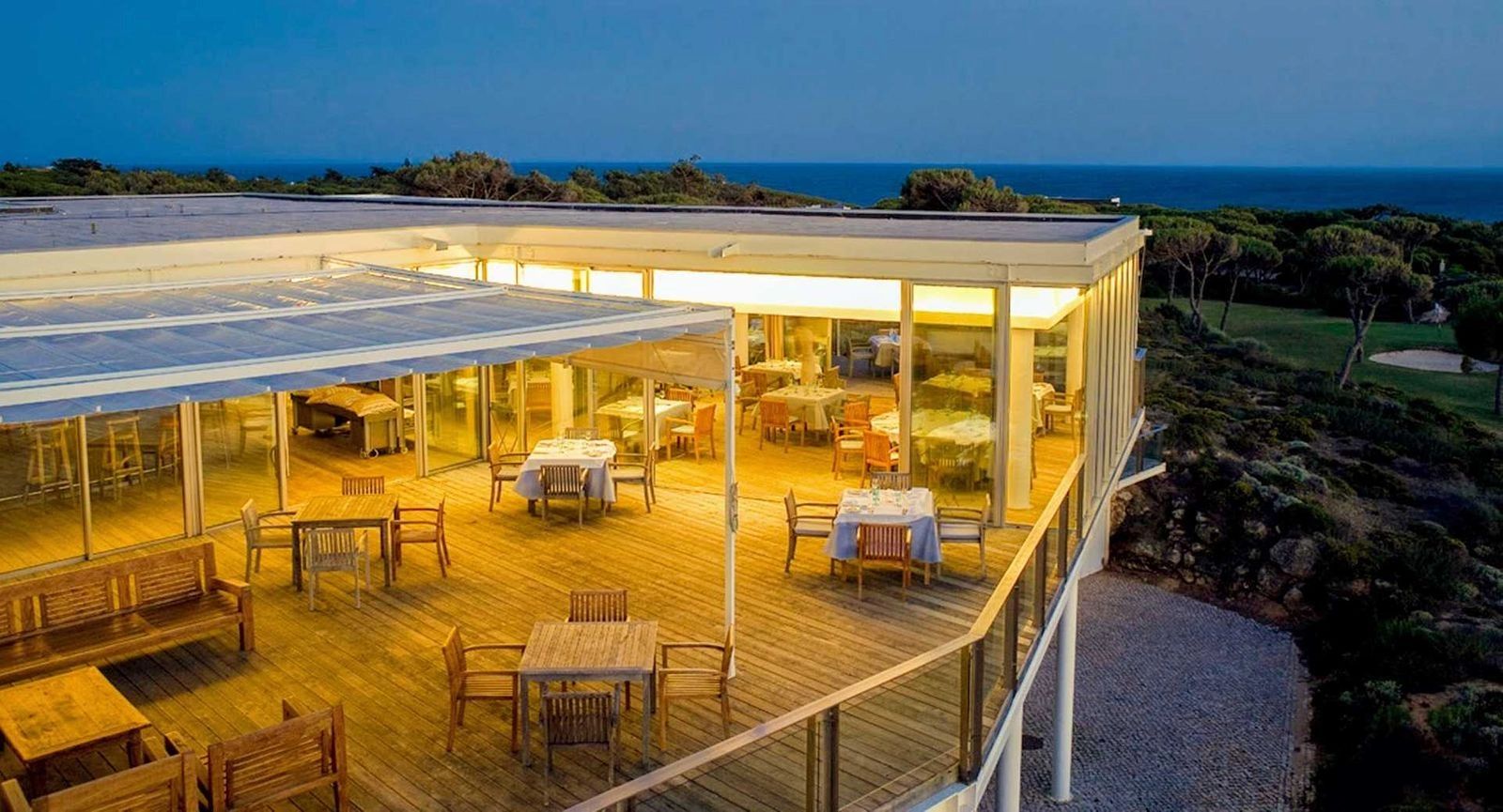 A terrace of a hotel well-lit with yellow lights, with wooden tables and chairs on top and a view to the greenery outside