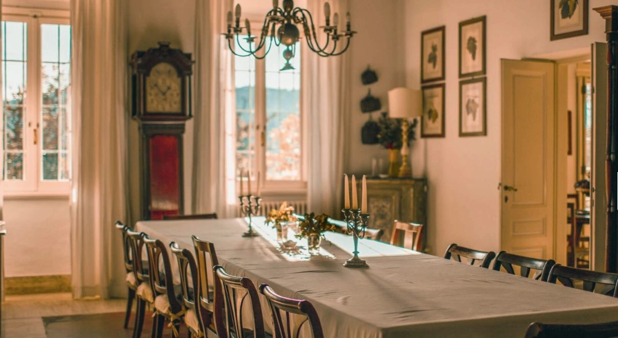 The villa dining room laid along one long table under a green iron chandelier, a red-cased longcase clock standing against the wall.