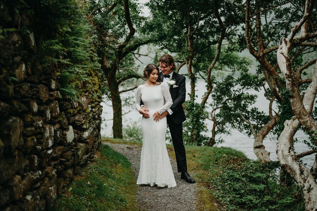 Groom hugging his bride from behind in a lush green nature spot with a lake in the background and tall trees at the shore