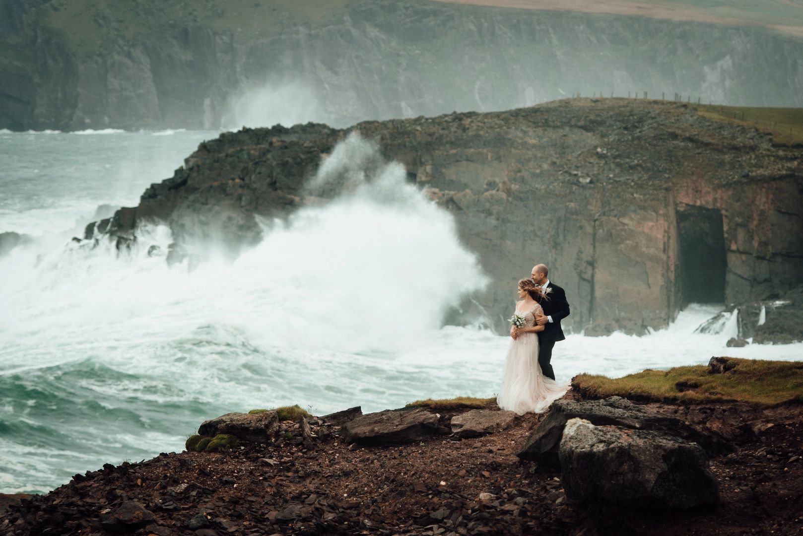 Newlyweds atop a rocky cliff overlooking Atlantic Ocean waves during their destination elopement in Ireland