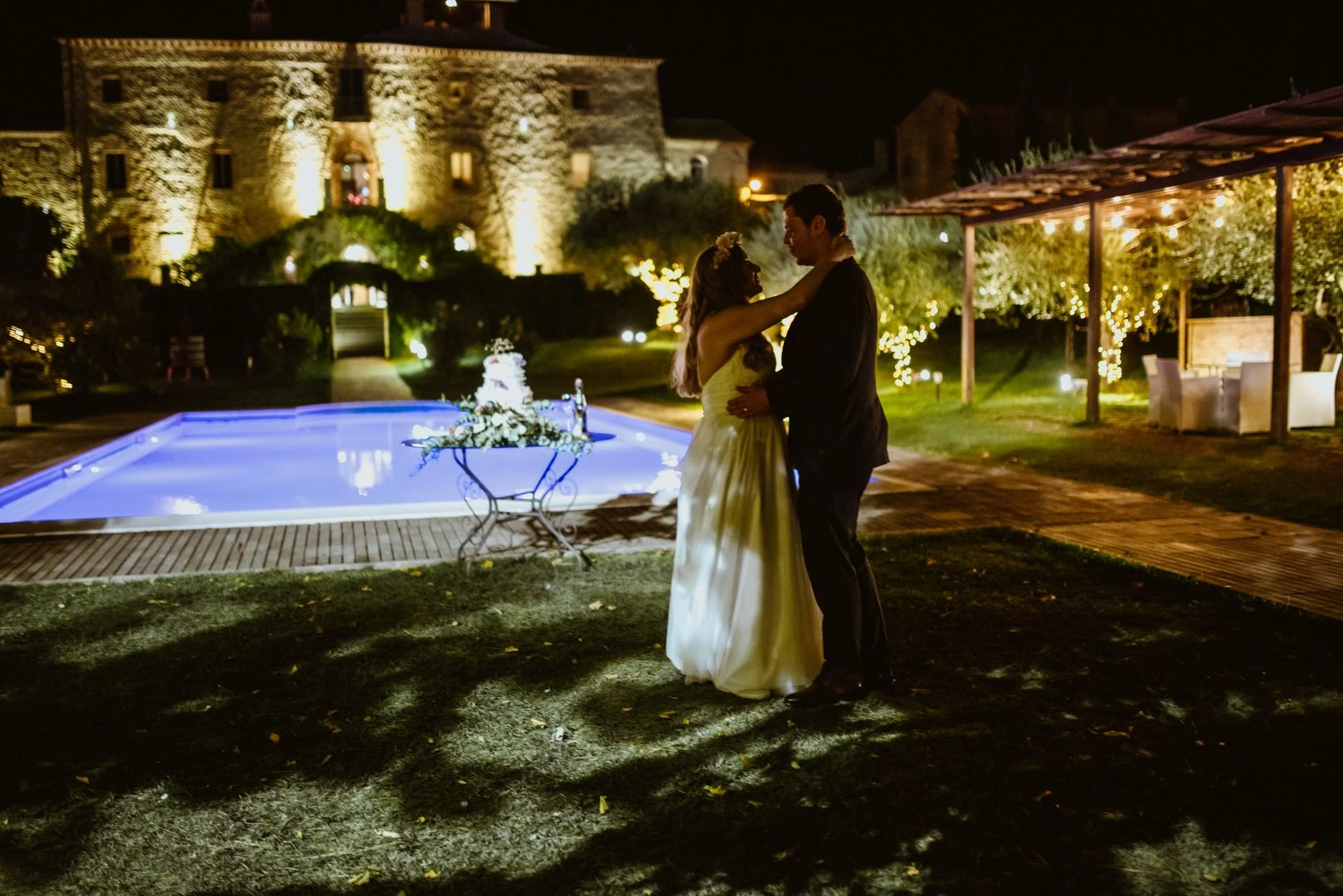 Bride and groom dancing at night with a pool, a villa, and their wedding cake in the background
