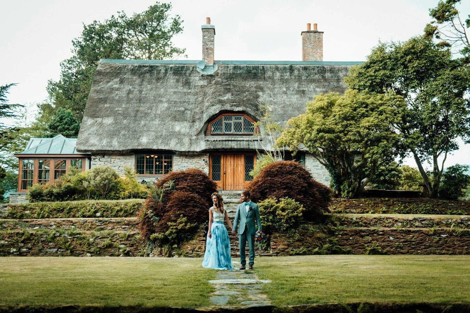 A thatched-roof cottage surrounded by trees and greenery, with a couple in front during their destination elopement in Ireland