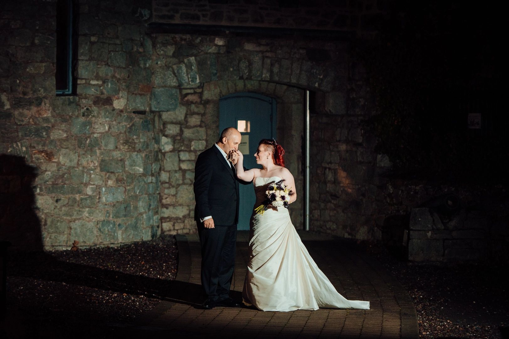 Groom kissing his bride's hand in front of a stone-walled castle at night