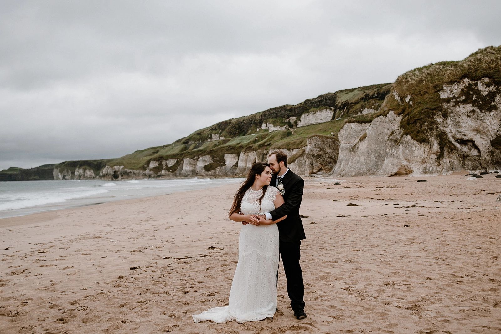 Groom hugging his bride from behind on white-sand beach with white rocks on the coast during their  elopement in Northern Ireland