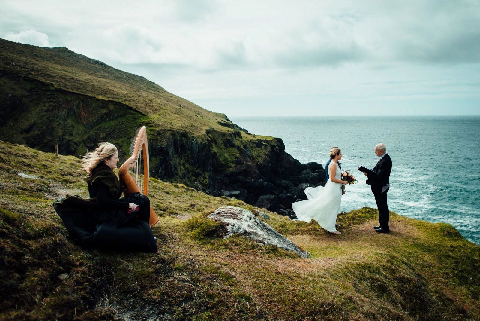 Clifftop elopement ceremony on the Dingle Peninsula: a harpist plays on the grass while the bride faces her celebrant at the edge, the Atlantic below.