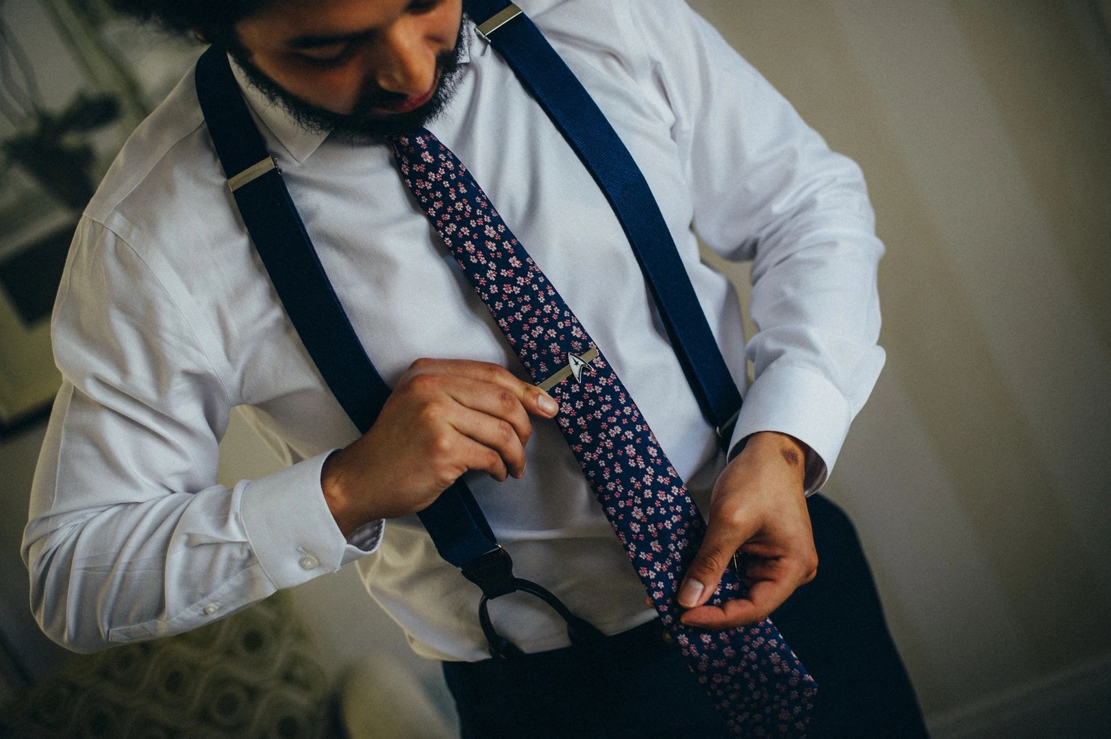 Groom in suspenders and black pants, fixing his floral tie
