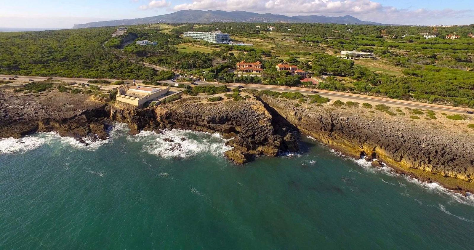 Hotels sitting in the middle of forested coastlands and the sea with waves splashing against the cliffs in Estoril, Portugal