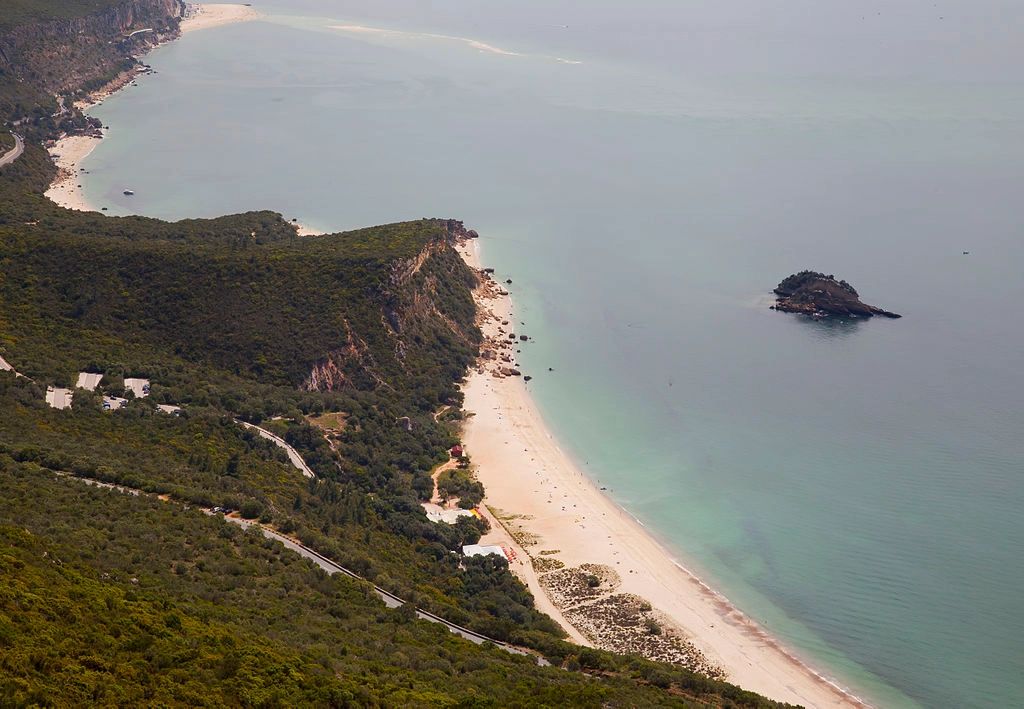 A white-sand beach next to the ocean with cliffs covered in lush greenery in Parque Natural da Arrábida, Portugal