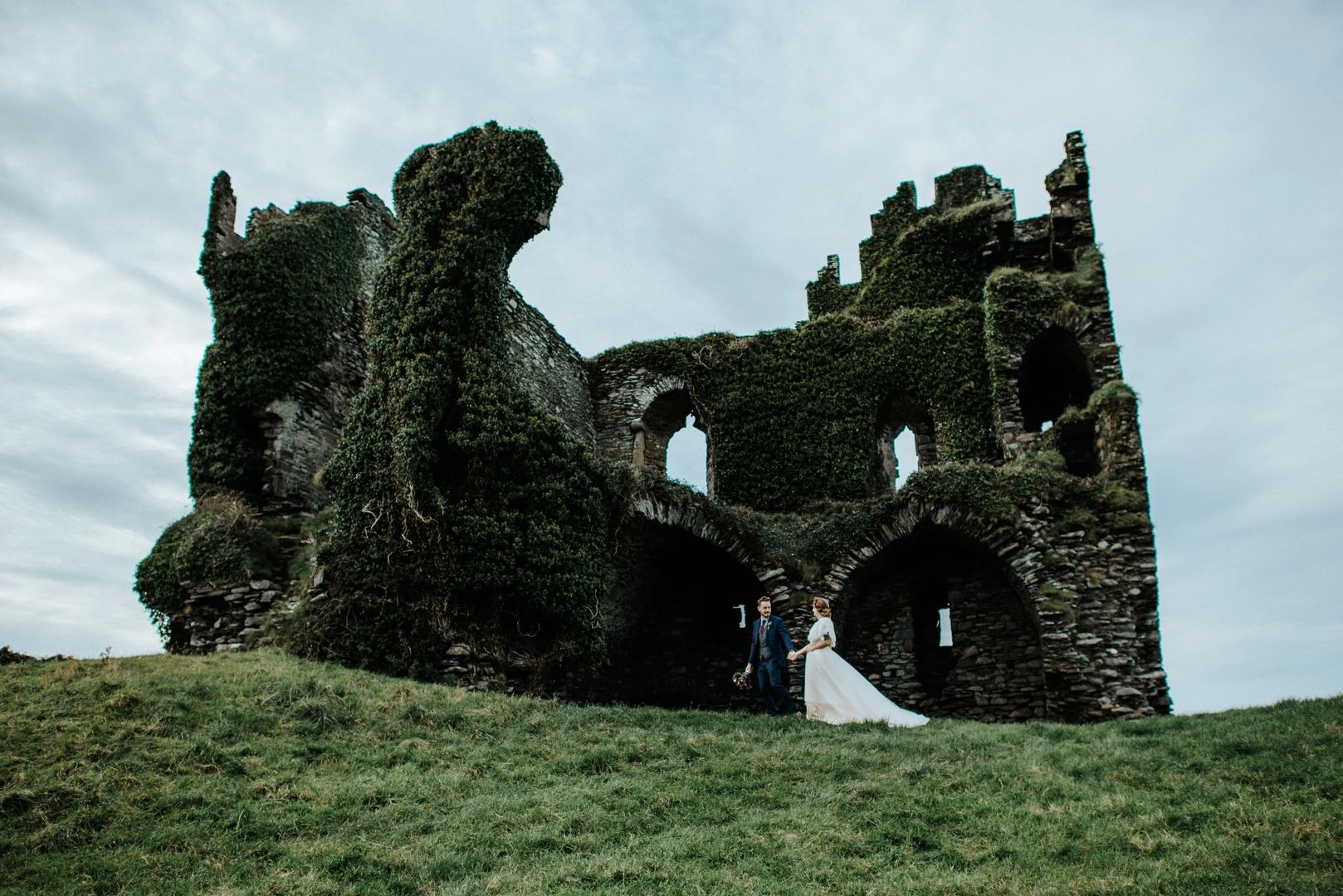 Bride and groom walking on a lush green field in front of Ballycarbery Castle for their destination wedding in Ireland