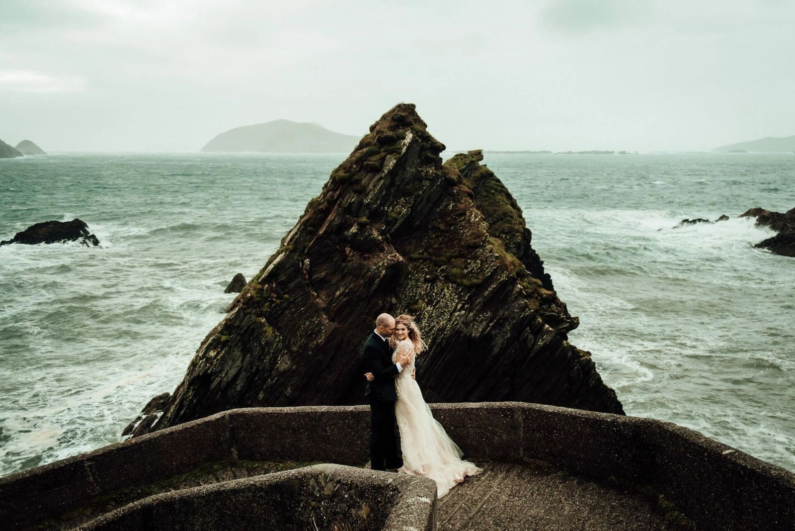 Slea Head, a clifftop elopement spot near Dingle, Ireland: newlyweds embrace on Dunquin Pier's stone-walled path above a pyramid sea stack.