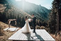 Couple holding hands on a wooden platform with mountains and alpine forests in the background at a wedding in Germany.