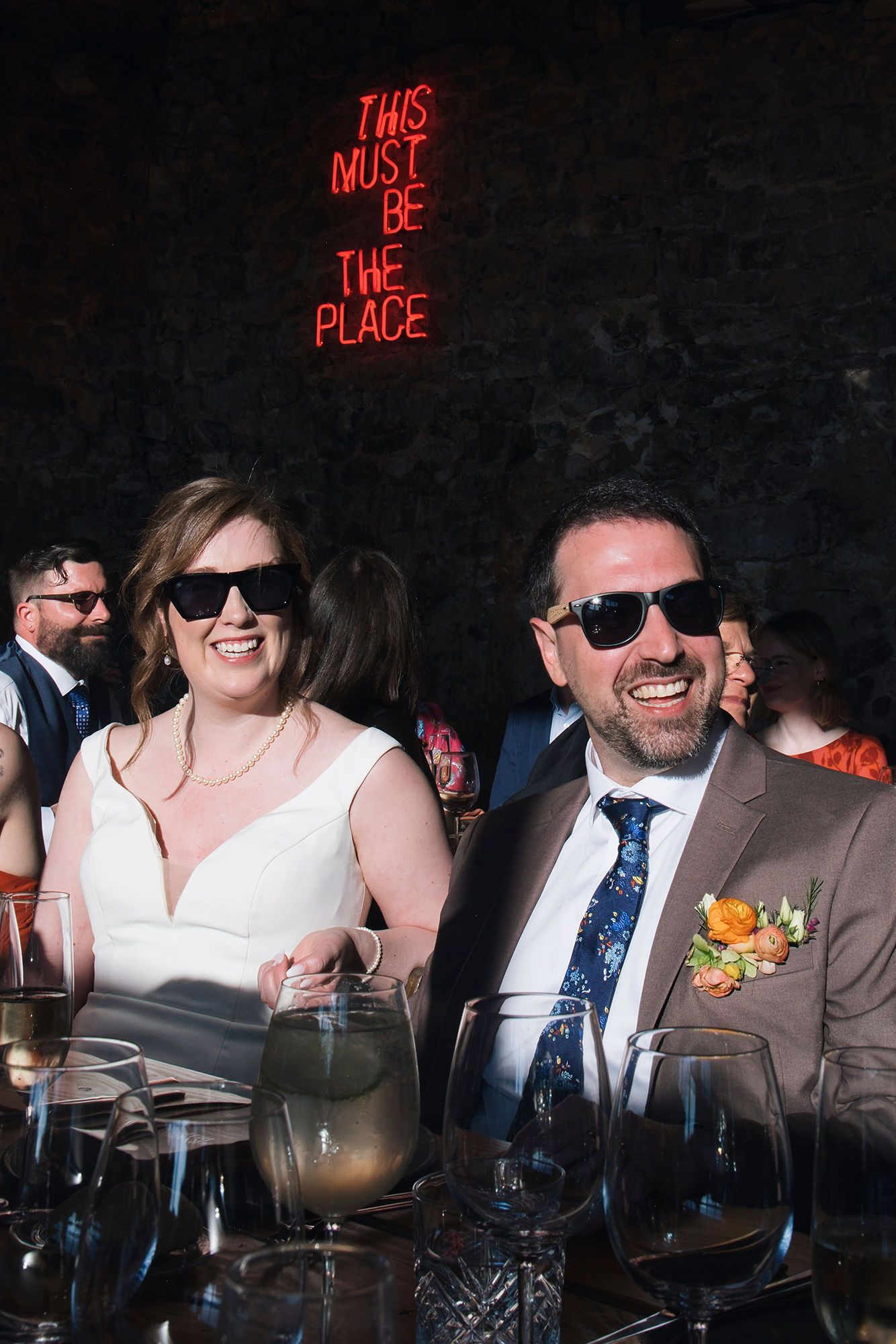 Bride and groom sit down, wearing sunglasses, watching, and smiling during the reception of their Spanish destination wedding