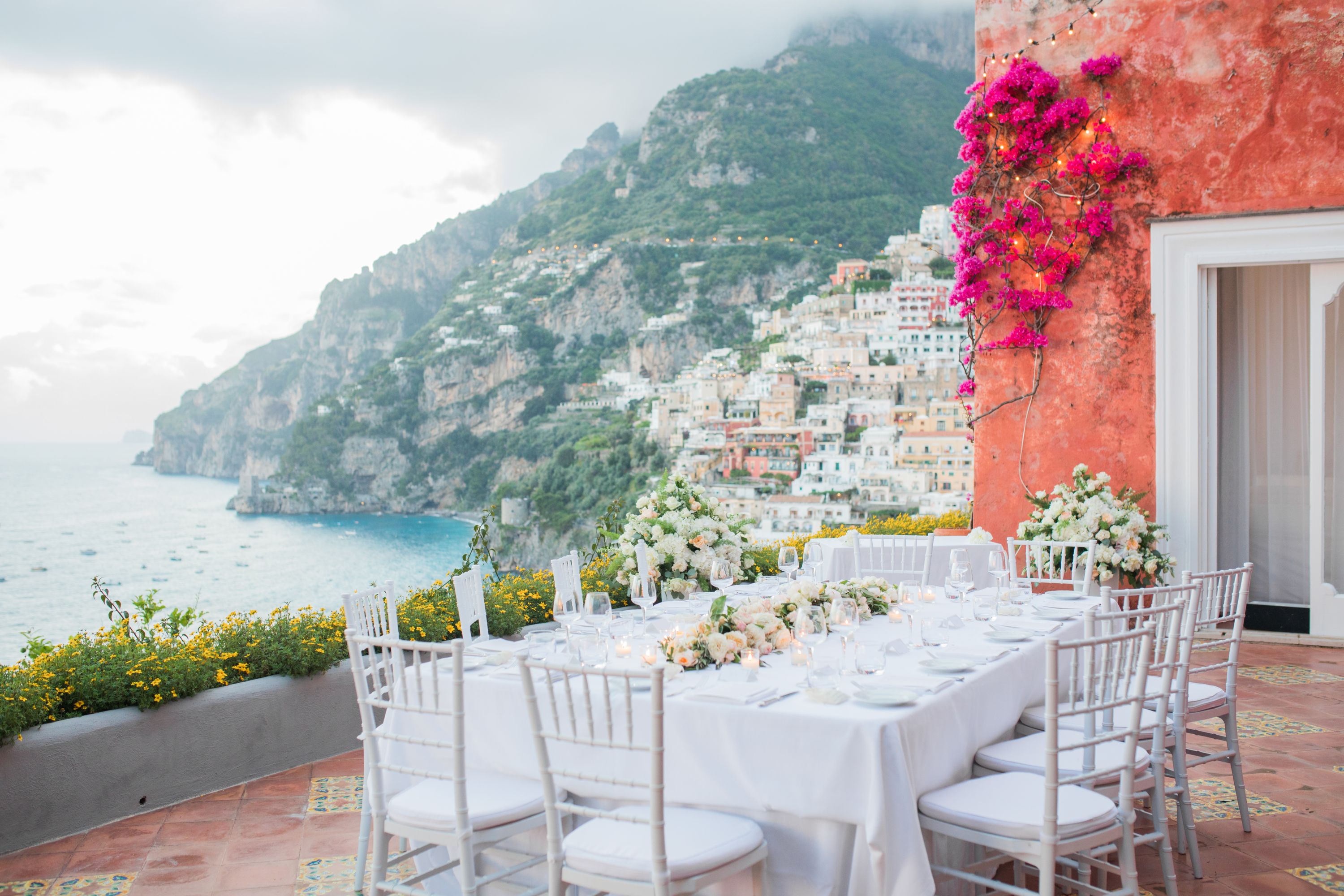 White tables and chairs arranged on top of a terrace of a hotel with the Amalfi Coast views in the background