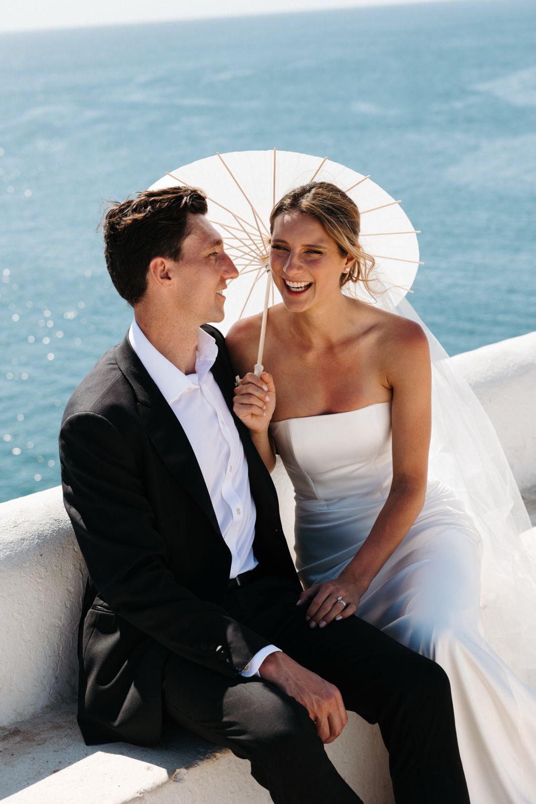 Bride laughing while holding a paper umbrella, with her groom and the sea in the background during their wedding in Portugal