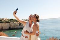 Two guests in a destination wedding in Portugal taking photos with cliffs and the ocean in the background.