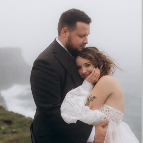 Bride buries her head into her groom’s chest while atop a cliff with ocean in the background during their Irish elopement