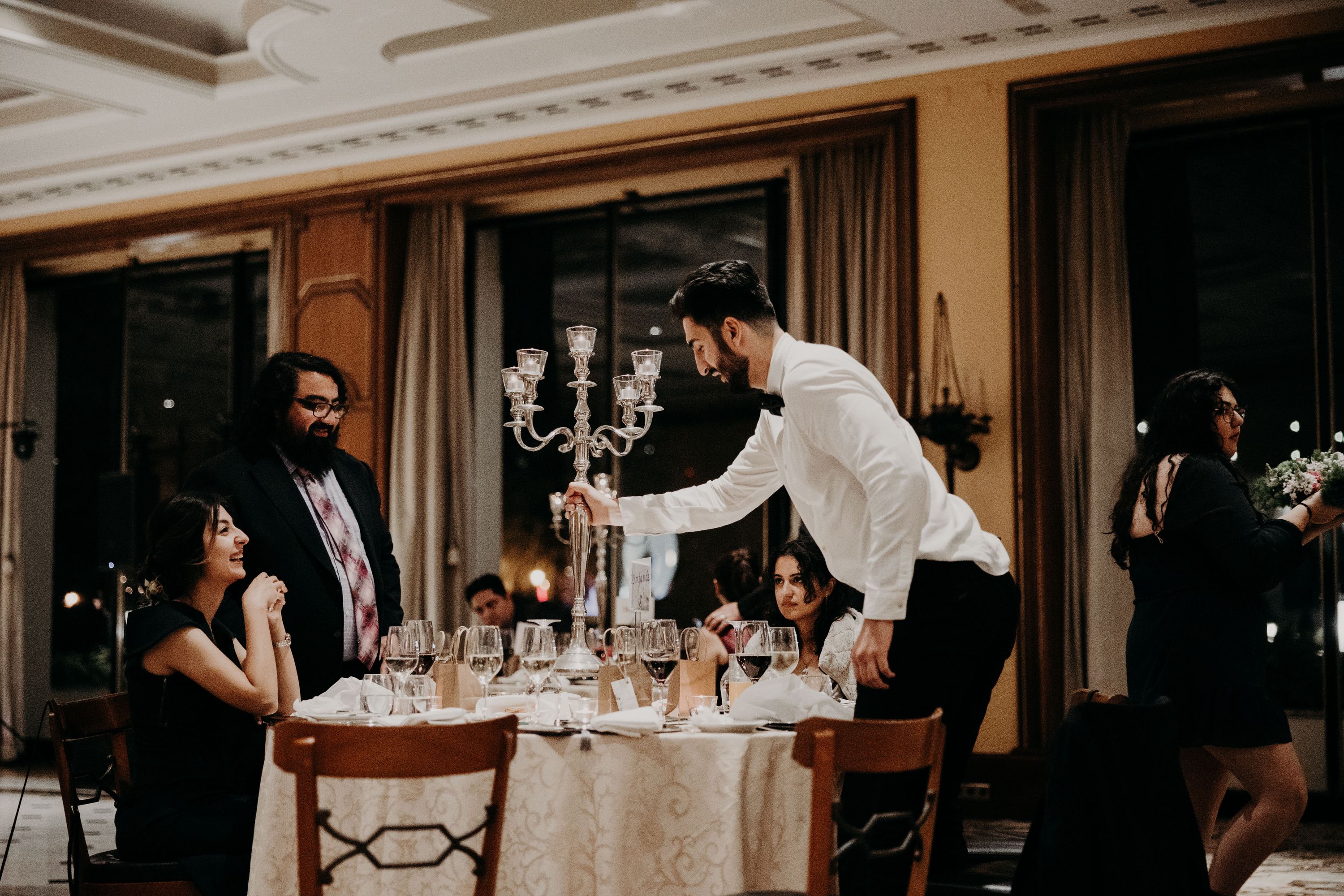 An event staff, wearing white long sleeves with a bow tie, assisting guests seated on a table during a wedding reception