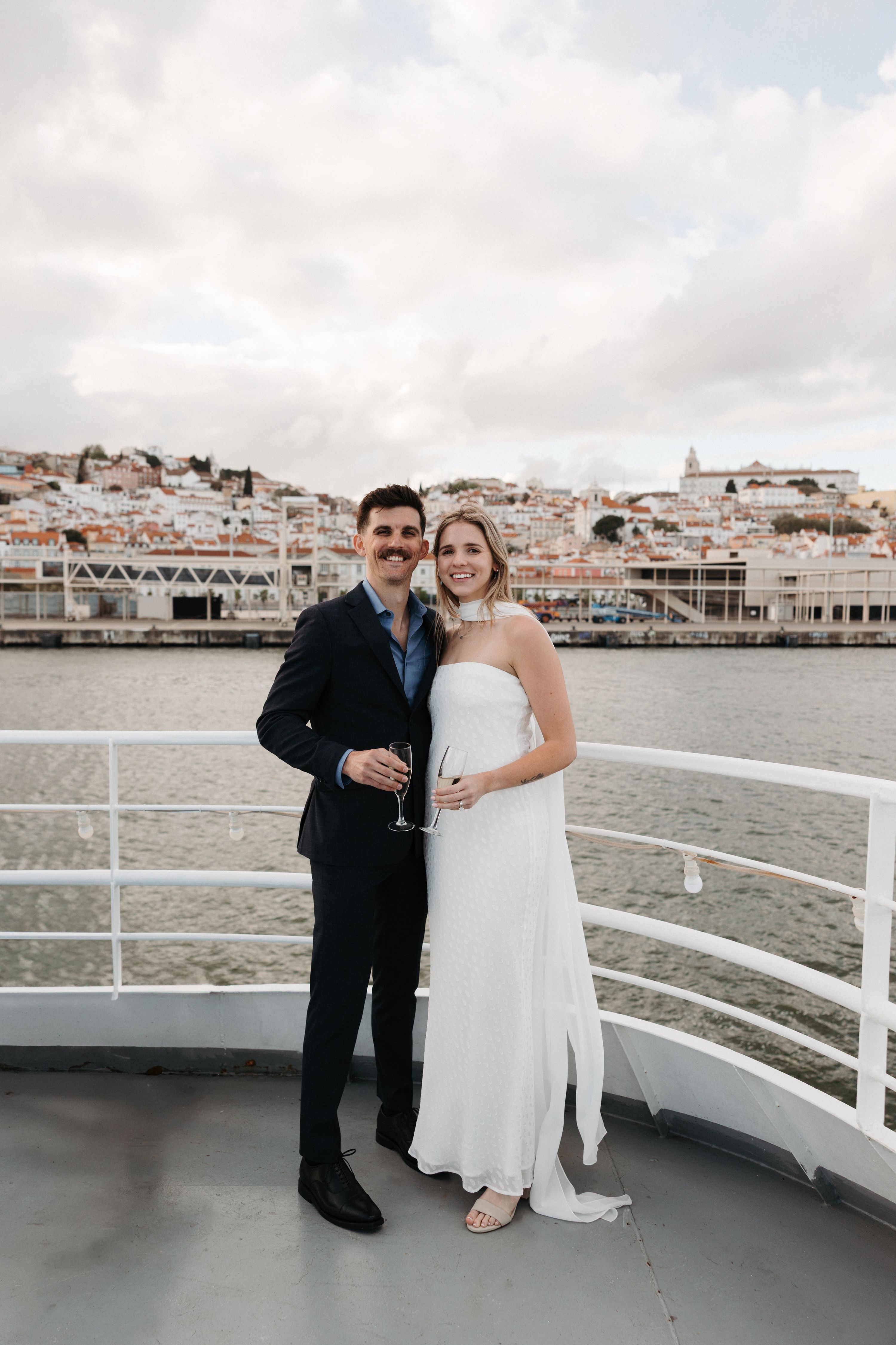 A couple holding champagne glasses at the edge of a boat with Lisbon waters and the Portuguese cityscape in the background