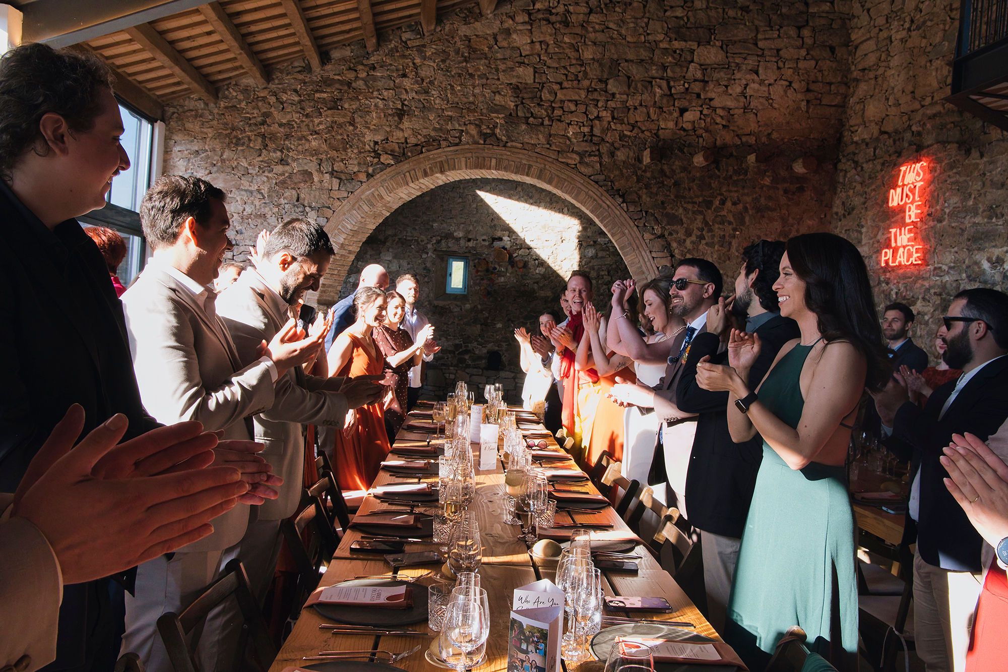 Guests standing up and clapping in front of a long table inside a medieval banquet hall during a wedding reception in Spain