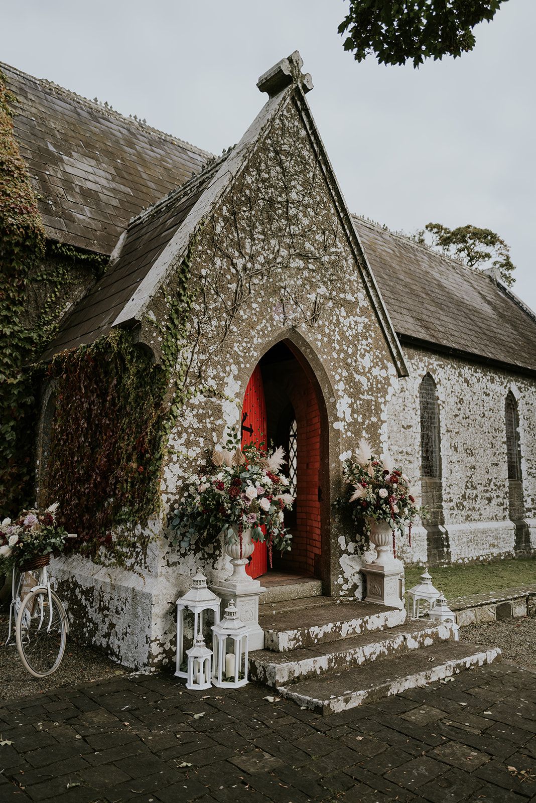 The south porch of St. John's Church, Knockainey, its red door open behind flower-filled urns and white lanterns, three stone steps up from the paving.