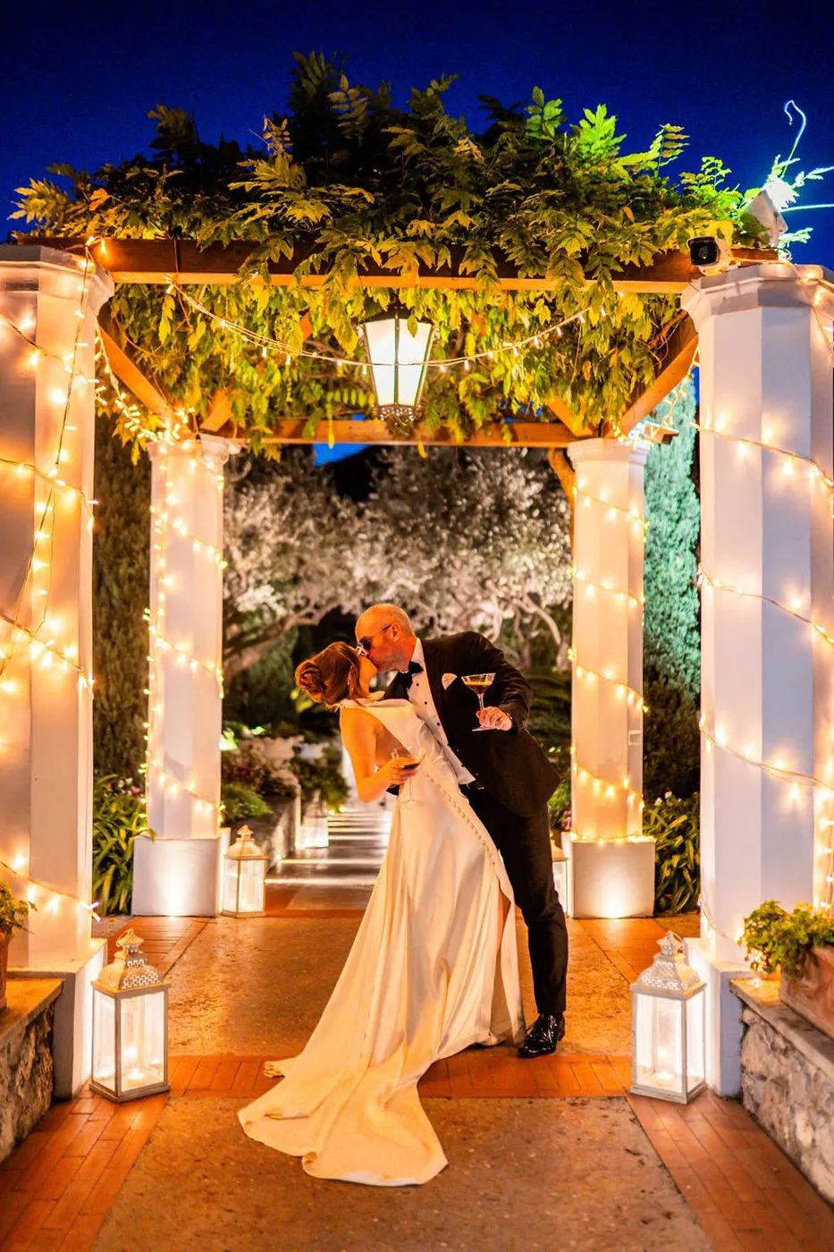Bride and groom kissing under a lighted gazebo with plants on the roof on the night of their vow renewal in Italy