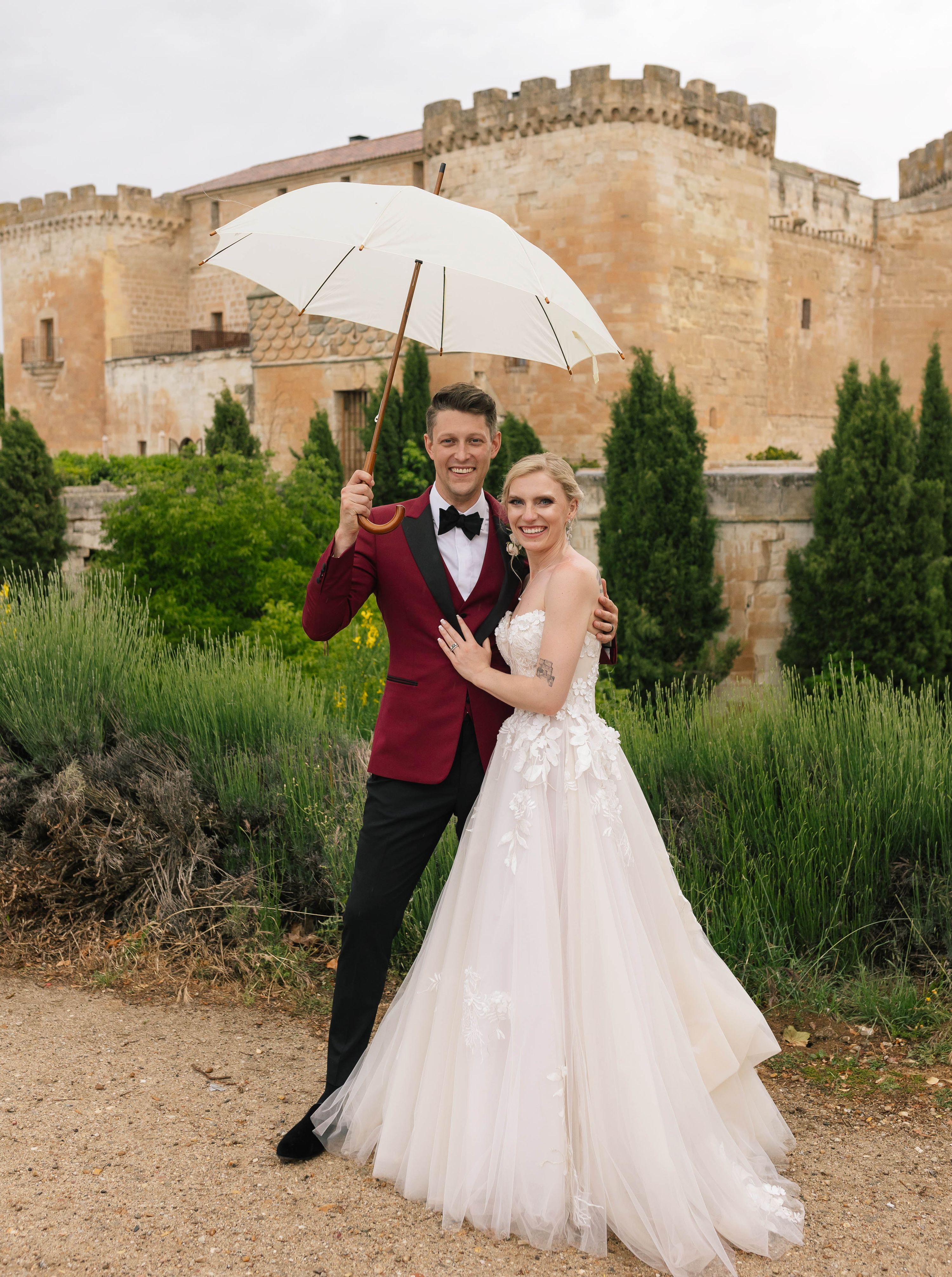 Newlyweds smile while looking at the camera, with a historic Spanish castle in the background, the groom holding an umbrella