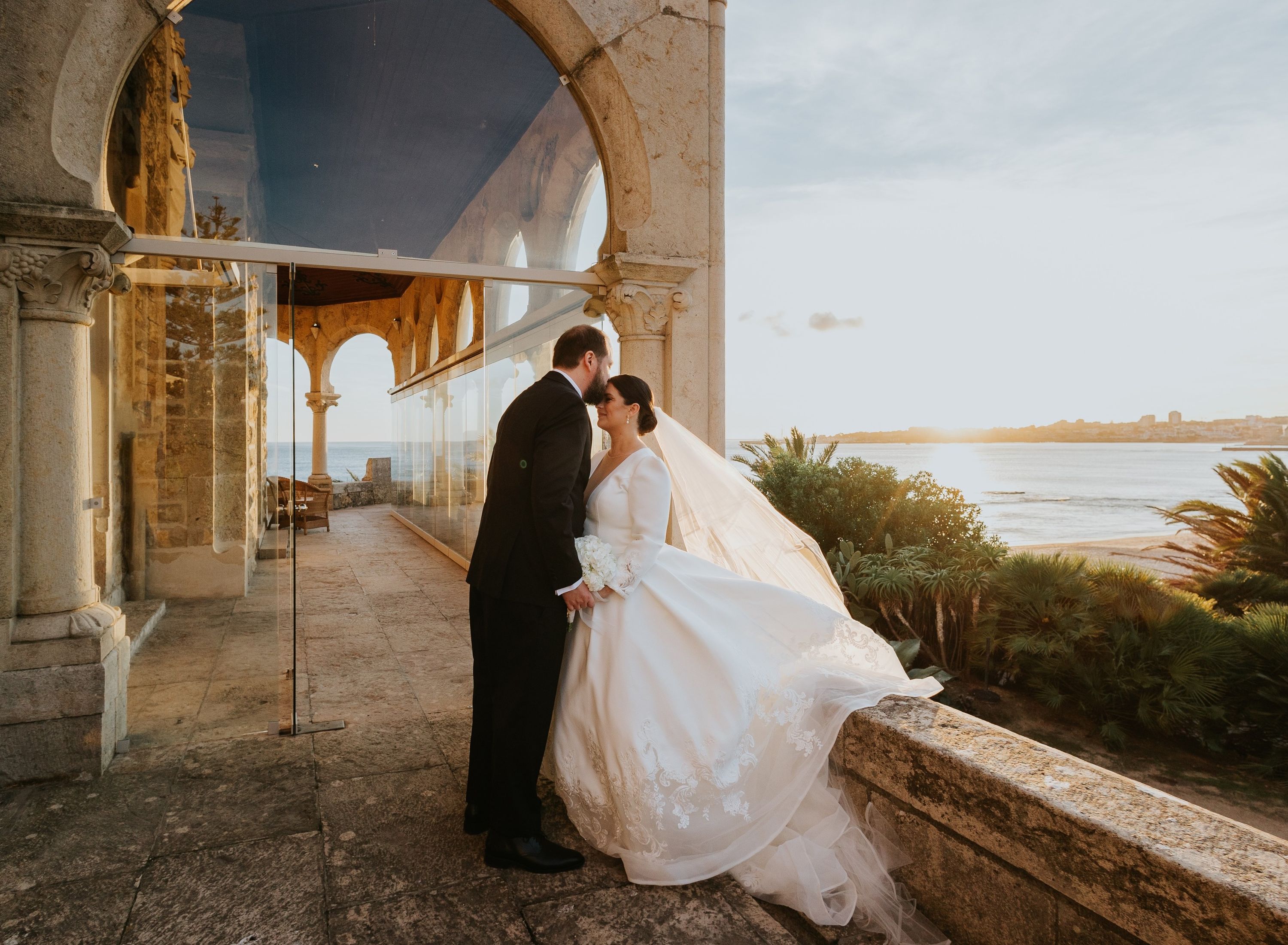 Groom kissing his bride’s forehead at a balcony of a historic castle overlooking the ocean