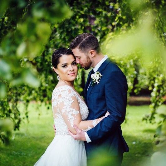 Bride looks at the camera while her groom smells her temple as they hug, with lush green landscapes of Ireland in the background