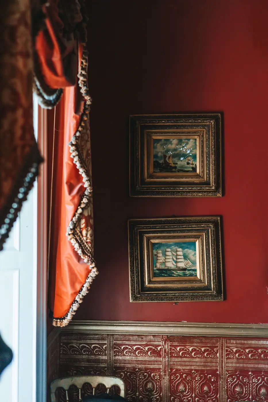 Red-walled dining room with gilt-framed paintings of sailing ships and tasselled orange curtains.