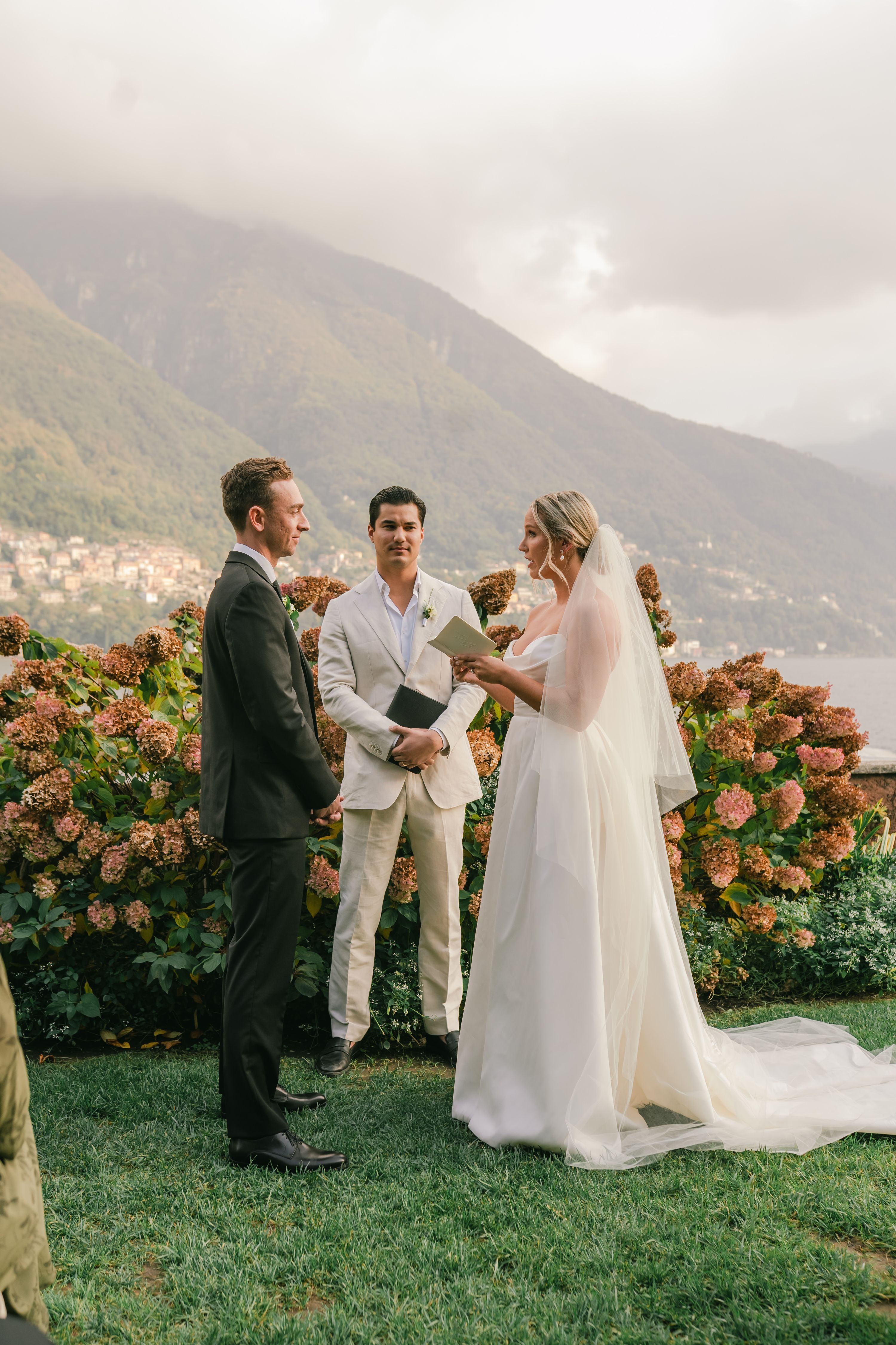 Bride recites vows before her groom with lush peach flowers and Lake Como in the background during their vow renewal in Italy
