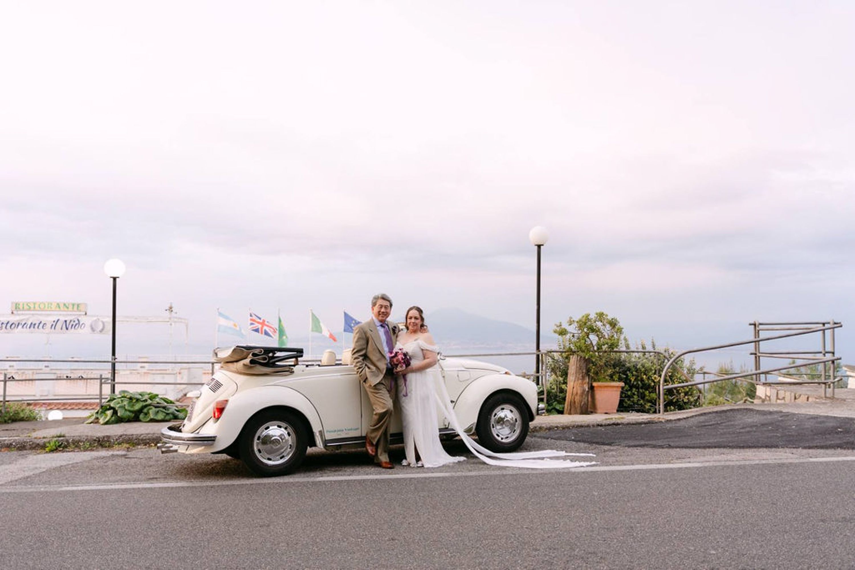 Newlyweds pose beside a classic convertible with coastal scenery at a small wedding in Italy