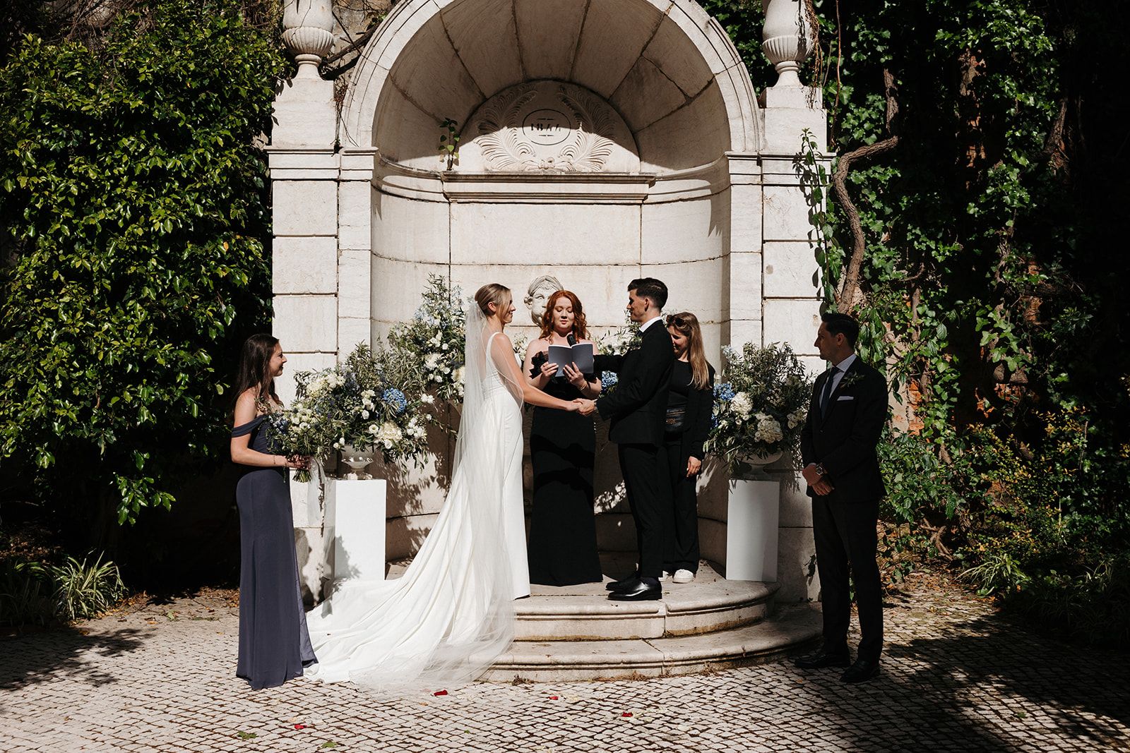 Groom hugs bride from behind in a beach setting with white sand and a portion of beach rocks behind