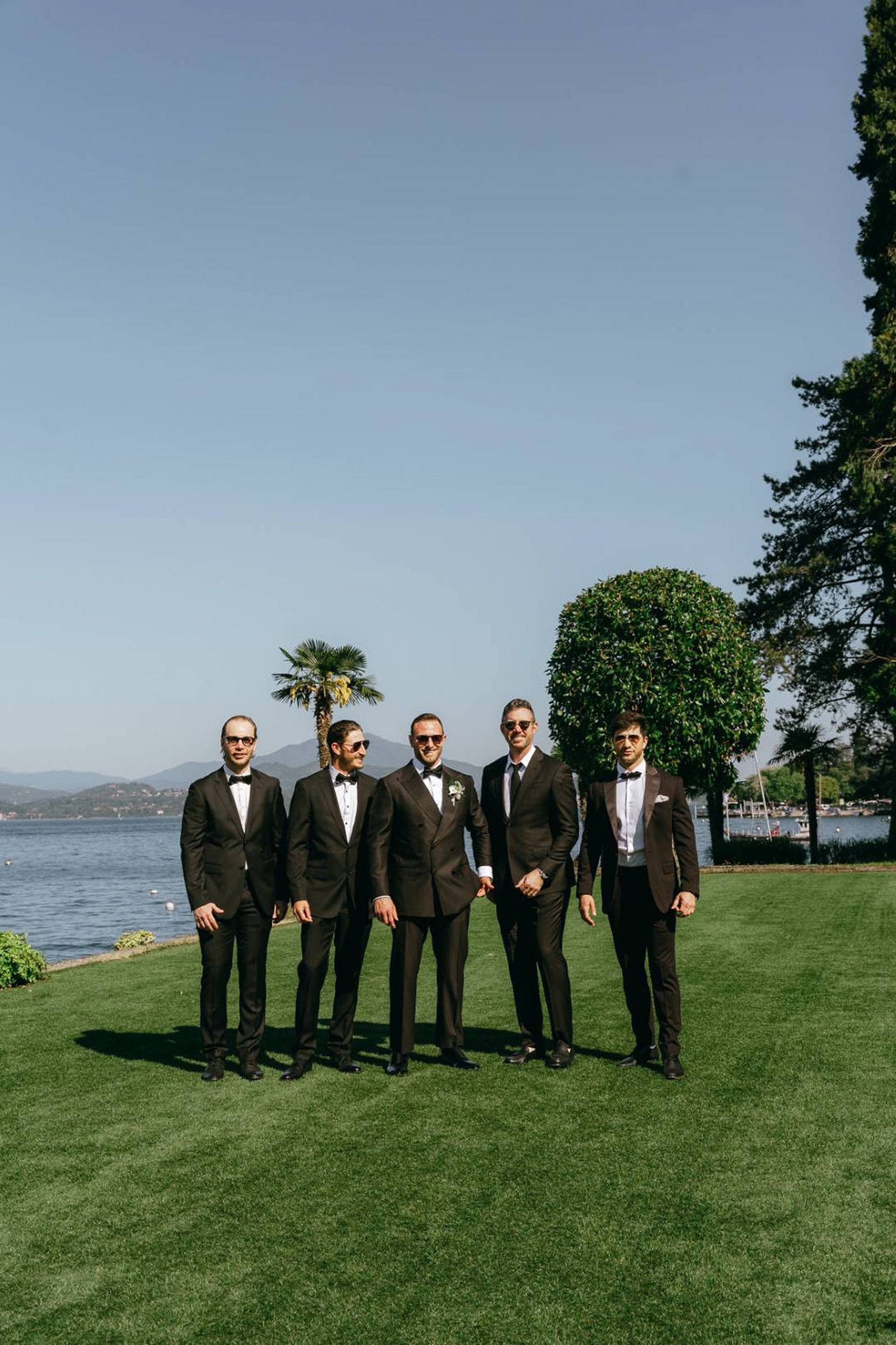 Groom and groomsmen posing by a lakeside lawn with mountain views at a small wedding in Italy
