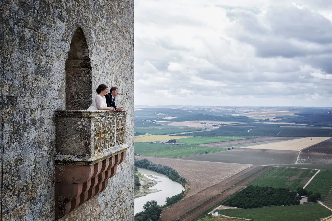 Couple standing on a castle terrace looks at the Andalusian landscape consisting of vineyards and wide open green spaces