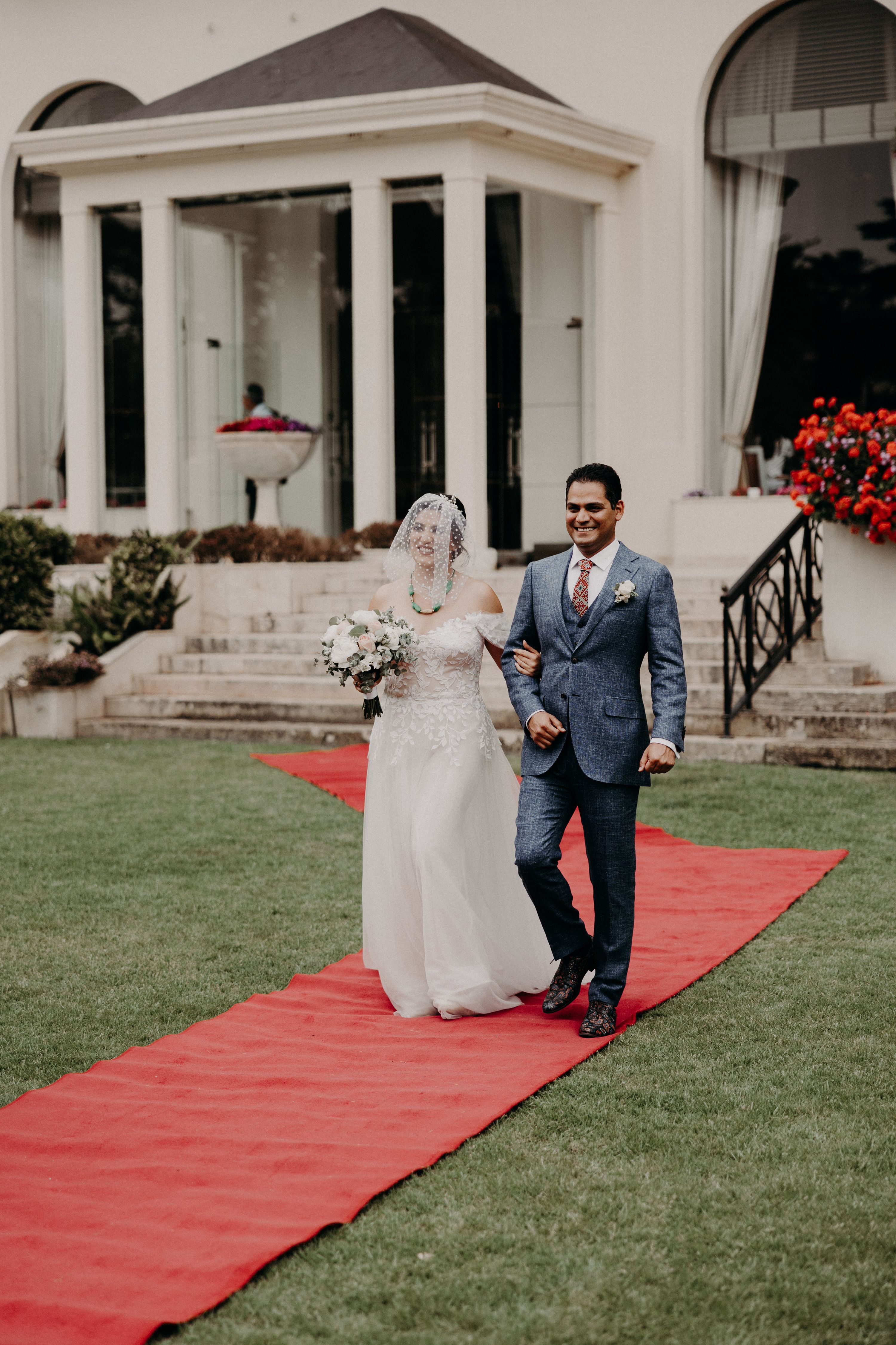 Bride and groom holding hands while walking down a red-carpet aisle with a white palace hotel in the background