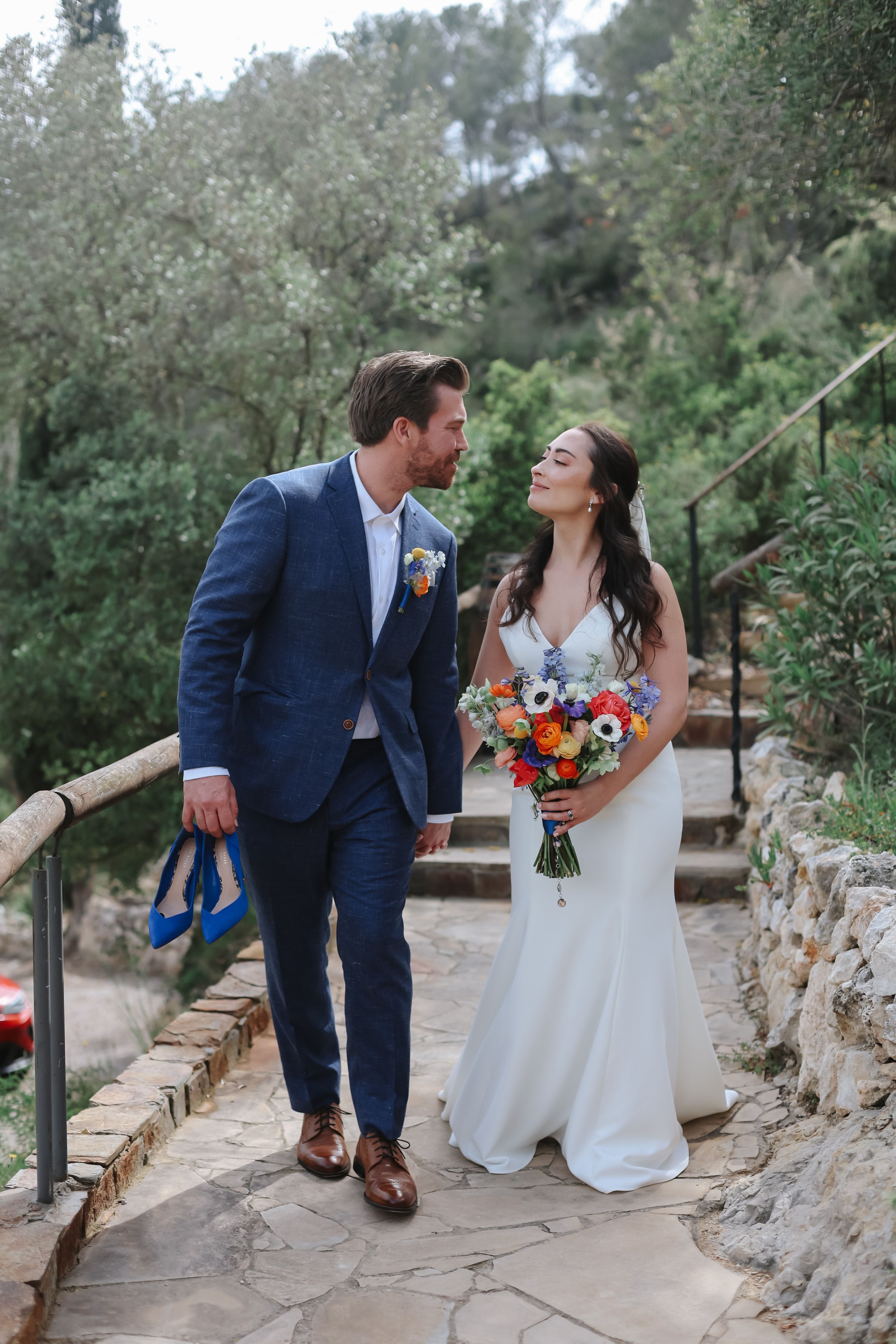 Bride and groom look at each other while the bride holds a colorful bouquet in her hands and lush greens in the background