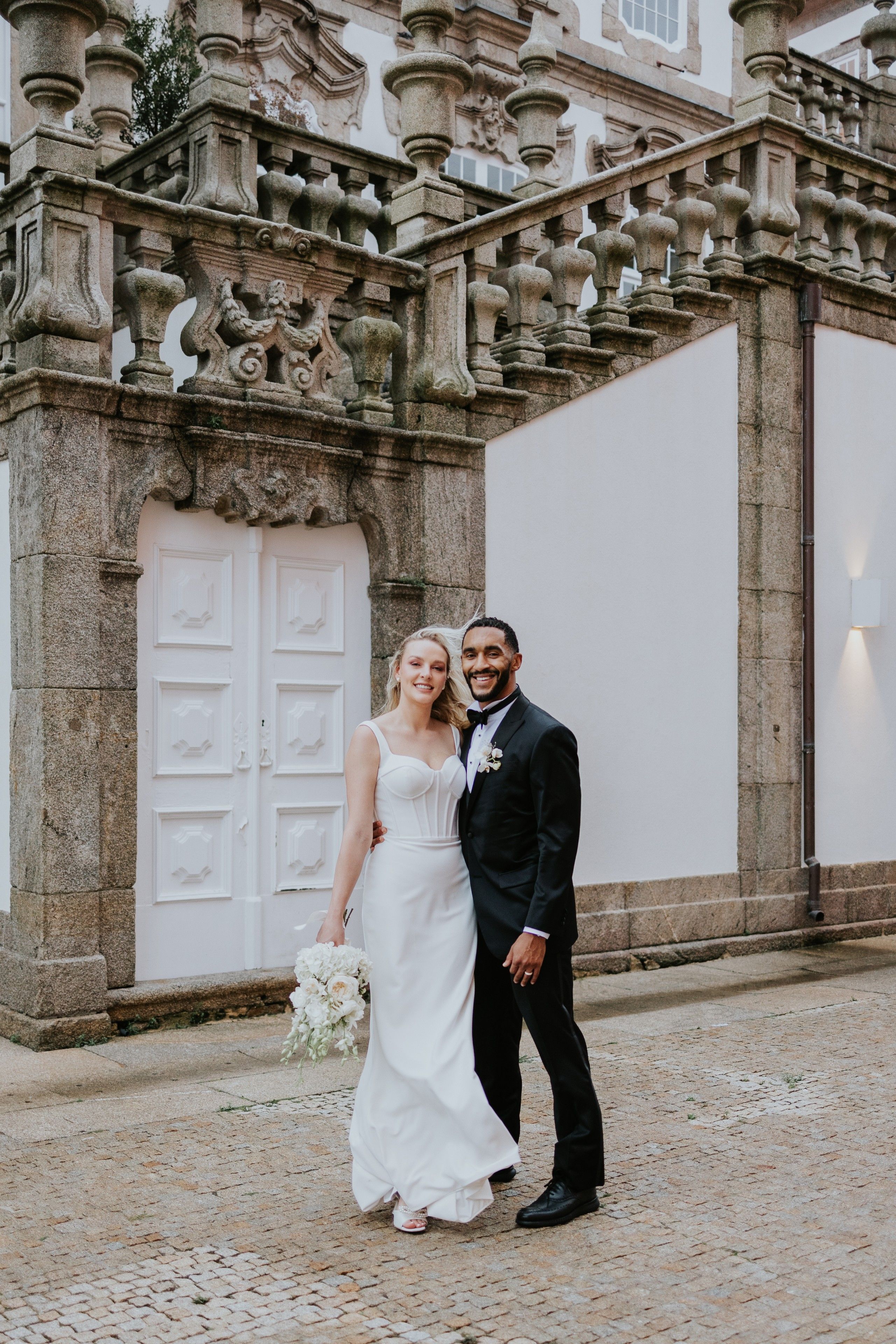 Newlyweds smiling at the camera while outdoors, with the wall of the castle in the background