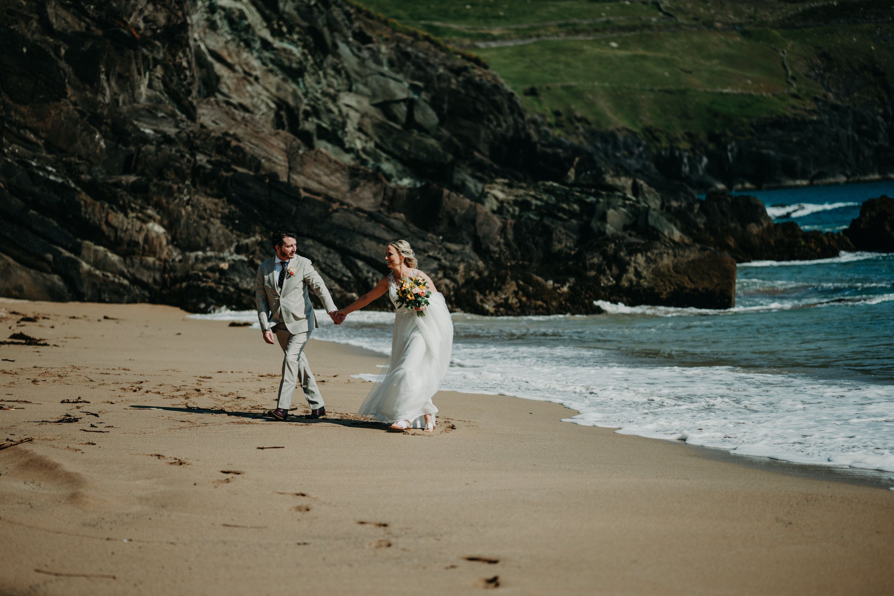 Bride with a yellow bouquet and groom in a pale suit stroll along Coumeenoole Beach near Slea Head, dark cliffs and surf behind.