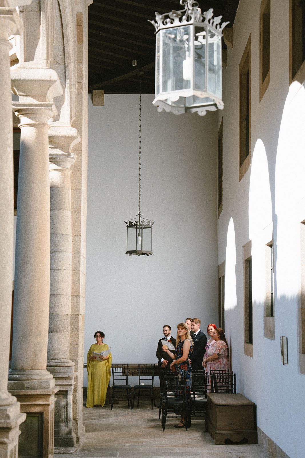 Wedding guests wait in a cloister gallery under hanging iron lanterns while the celebrant in yellow reads, Parador de Santiago.