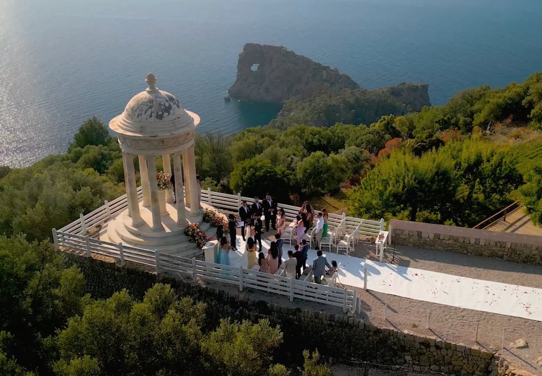 An outdoor wedding with a white stone gazebo in front atop a cliff overlooking the sea and the lush landscapes of Ibiza