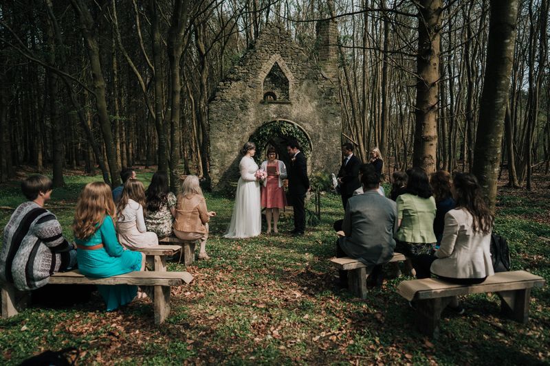 Bride and groom having an outdoor ceremony in a forest with a church ruin in the background during their Irish elopement