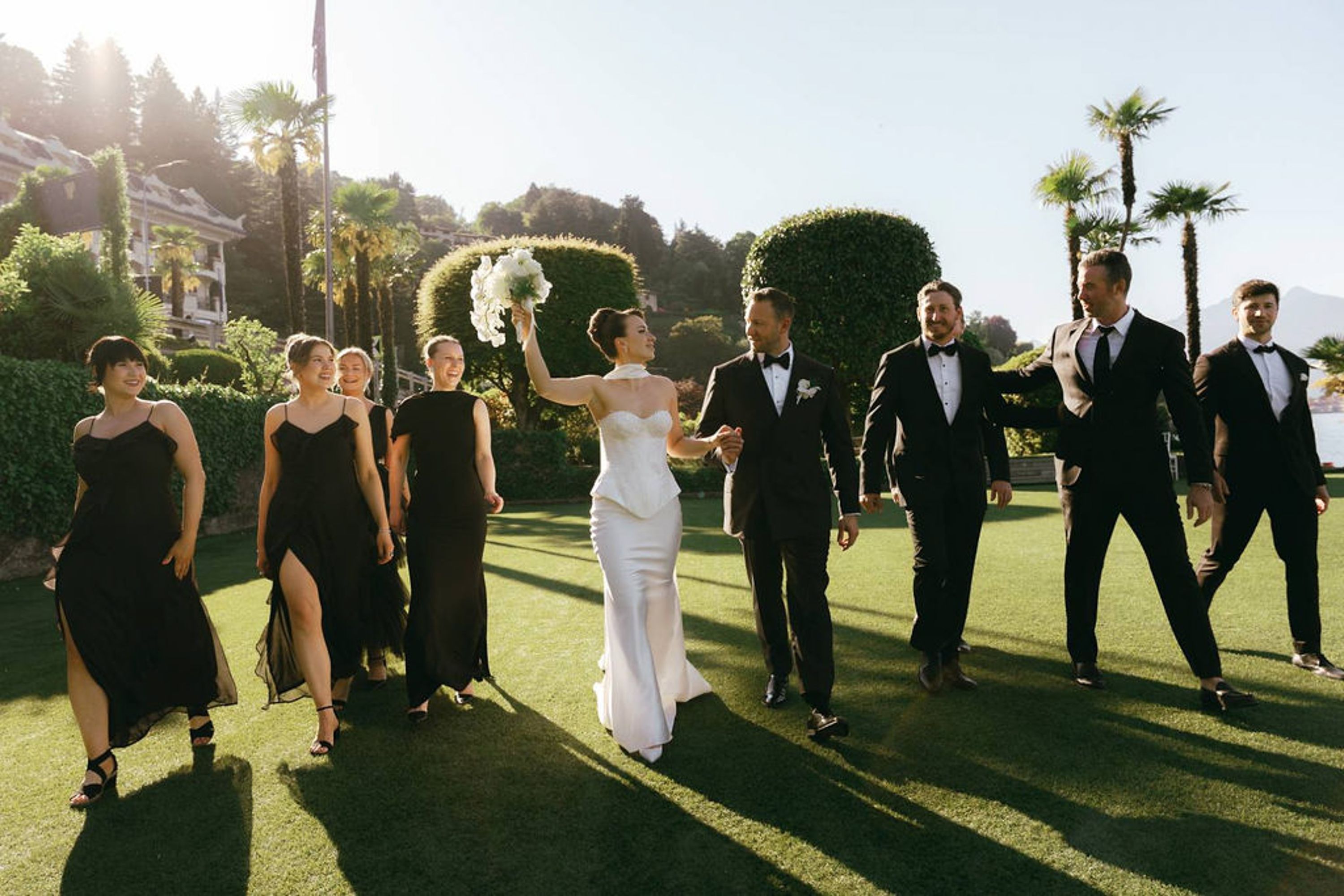 Bride and groom walking with their wedding party across sunlit gardens at a small wedding in Italy