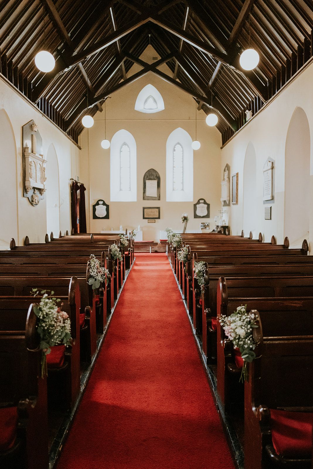 Looking down the red-carpeted aisle of St. John's Church, Knockainey, past pews tied with gypsophila to a ceremony table under twin lancet windows.