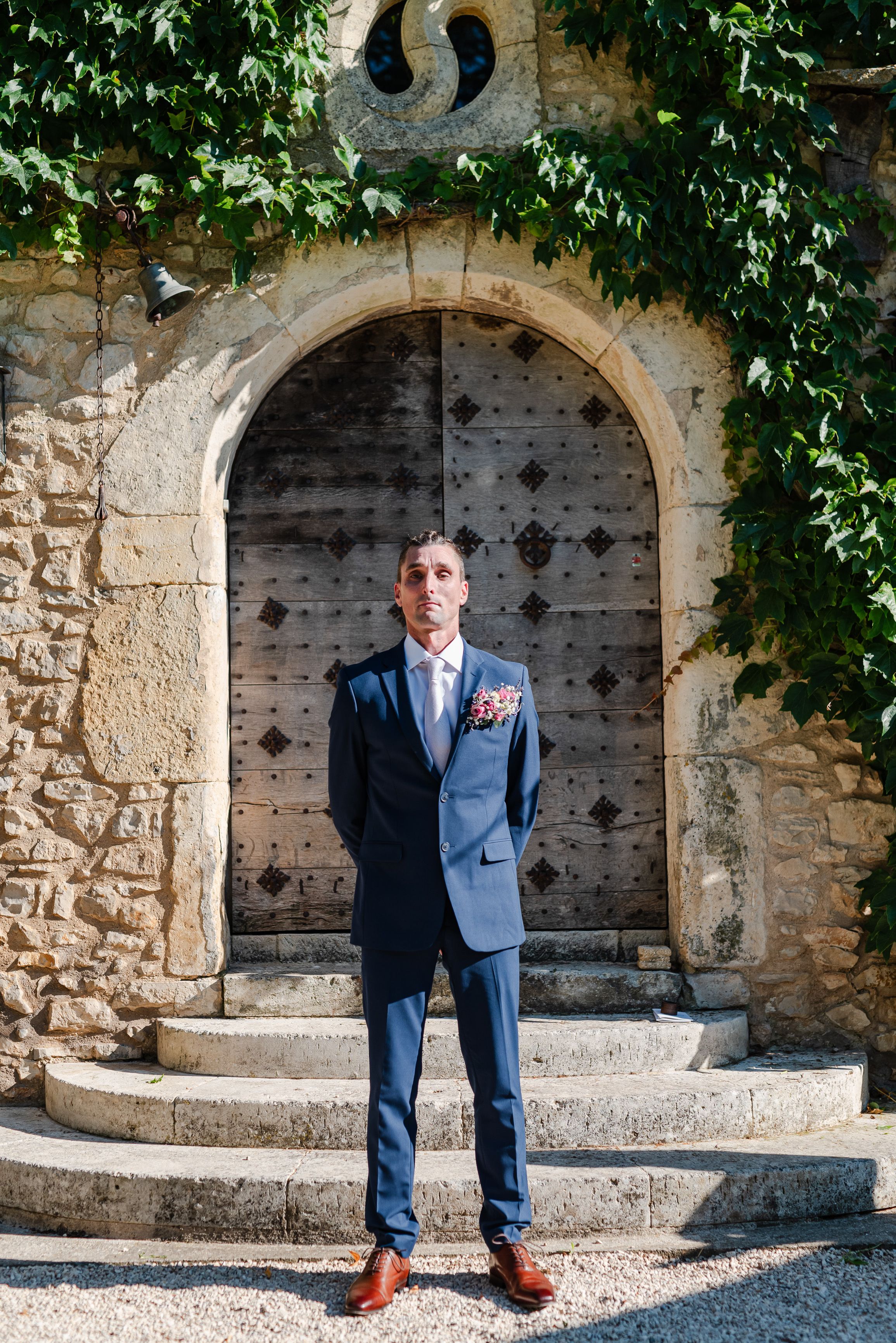 A groom wearing a blue suit standing near a stone archway during an intimate wedding in France